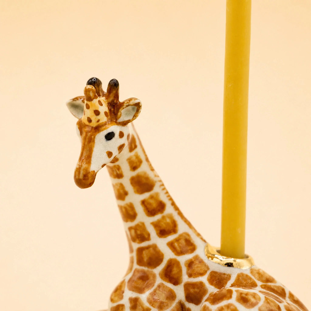 A close-up of the Giraffe Cake Topper — a handcrafted collectible ceramic piece featuring a yellow candle inserted on its back, set against a plain beige background.