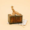 A Giraffe Cake Topper—crafted to evoke the charm of a collectible figurine—rests atop a vintage-style wooden box adorned with metal accents, reminiscent of a keepsake treasure chest. This arrangement is set on a star-patterned drawstring cloth bag, all displayed against a soft light beige background.