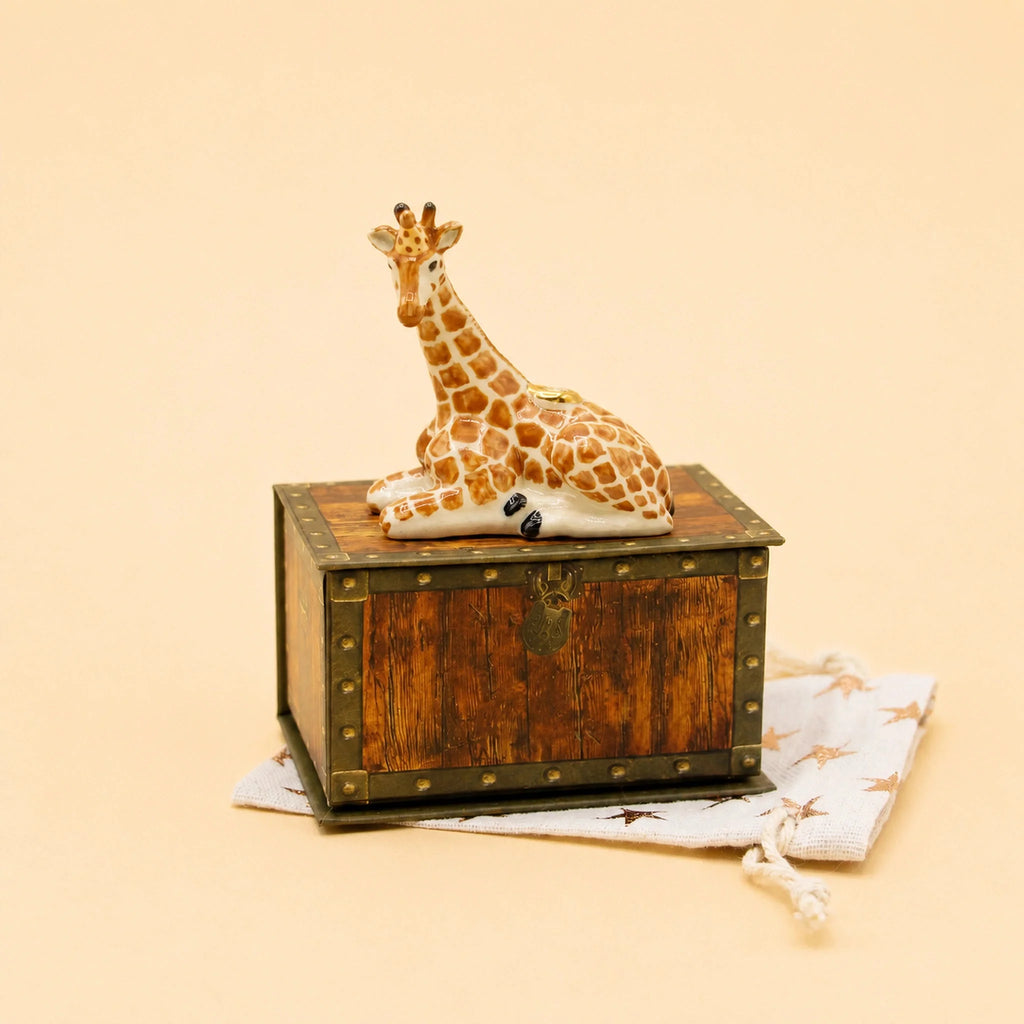 A Giraffe Cake Topper—crafted to evoke the charm of a collectible figurine—rests atop a vintage-style wooden box adorned with metal accents, reminiscent of a keepsake treasure chest. This arrangement is set on a star-patterned drawstring cloth bag, all displayed against a soft light beige background.