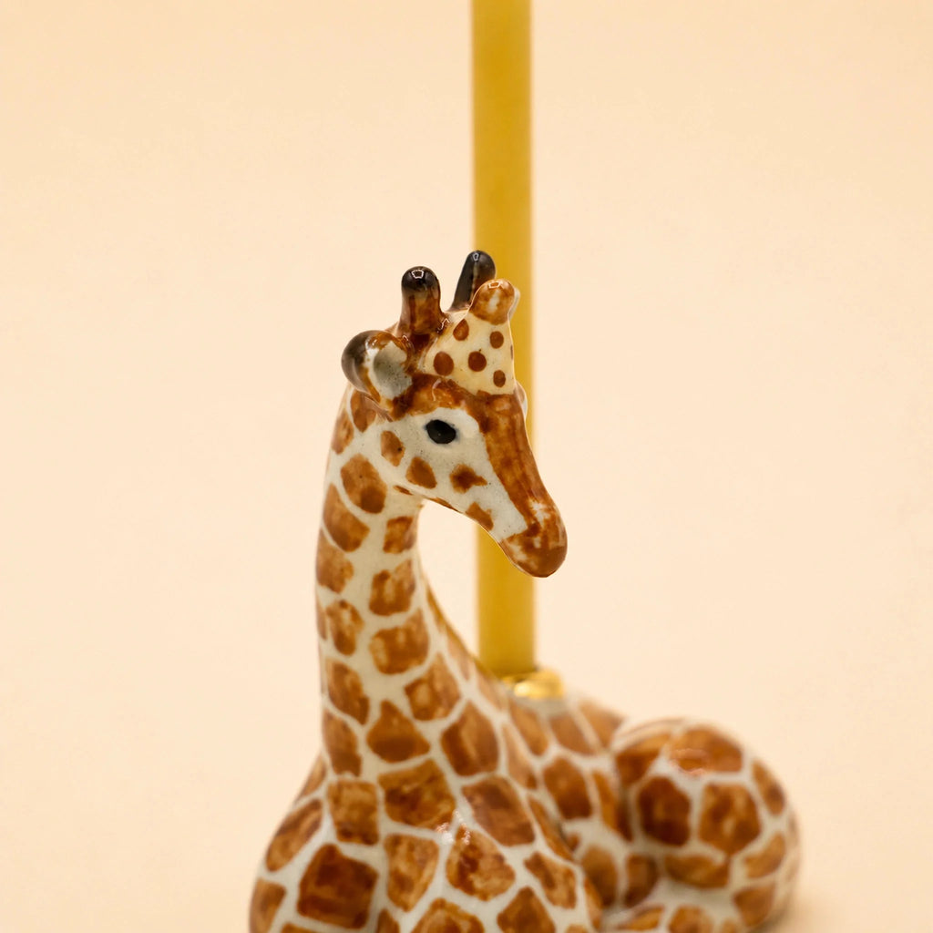 A handcrafted “Giraffe Cake Topper - Final Sale” ceramic figurine with brown spots is lying down, with a yellow candle positioned behind it against a plain, light beige background.