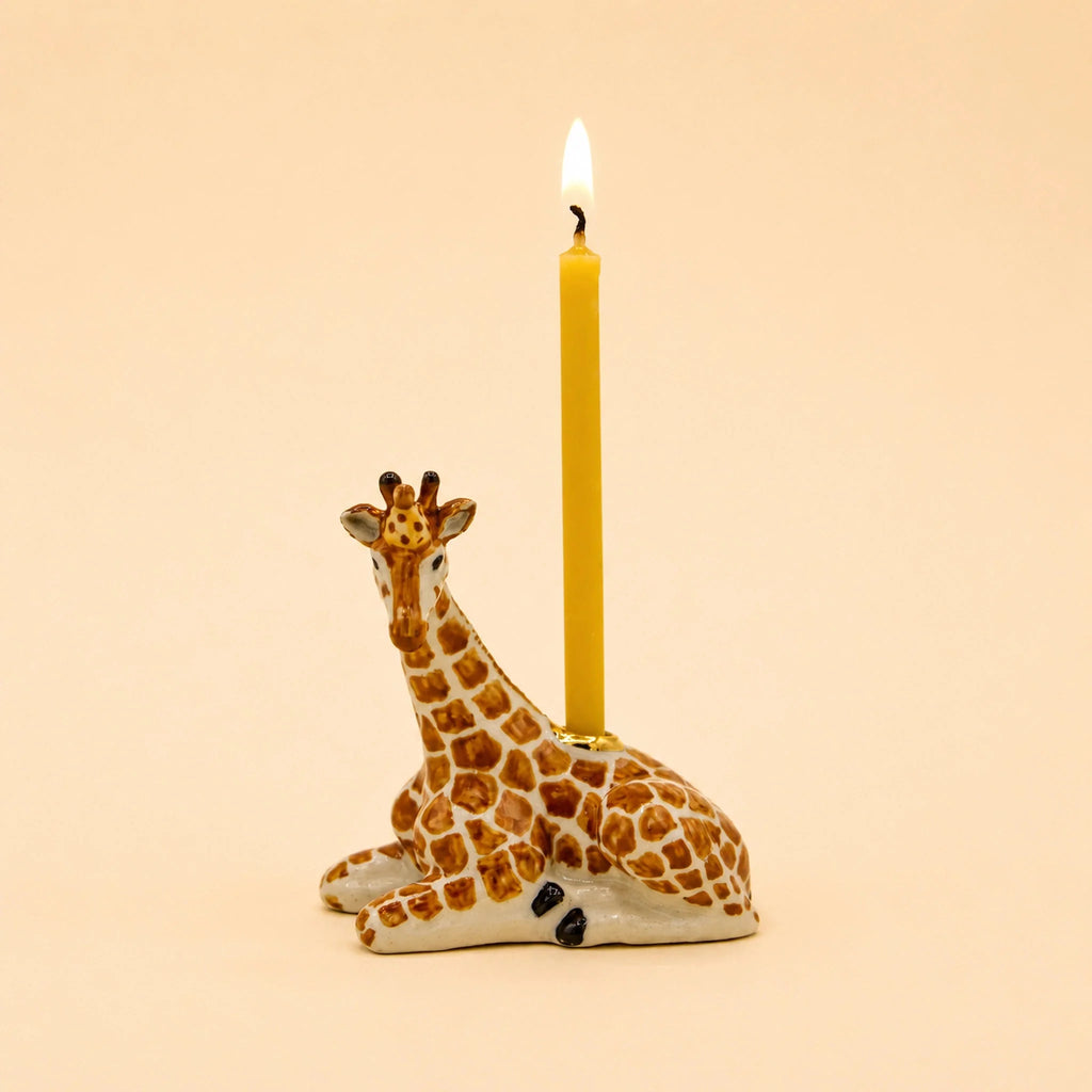 A yellow candle stands atop the Giraffe Cake Topper—a handcrafted, collectible ceramic holder shaped like a sitting giraffe—set against a plain beige background.