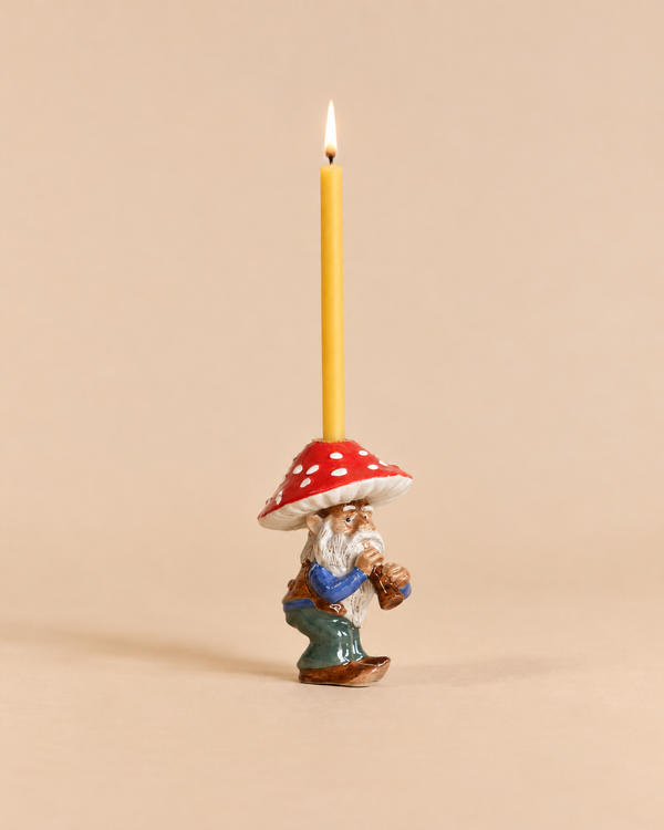 Add a whimsical touch to your space with the Mushroom Gnome Cake Topper—this heirloom-quality collectible features a ceramic gnome holding a lit yellow candle, dressed in a blue shirt and green pants with a red, white-spotted mushroom hat. Perfect as a keepsake or unique decor accent, this final sale item is photographed against a plain beige background.