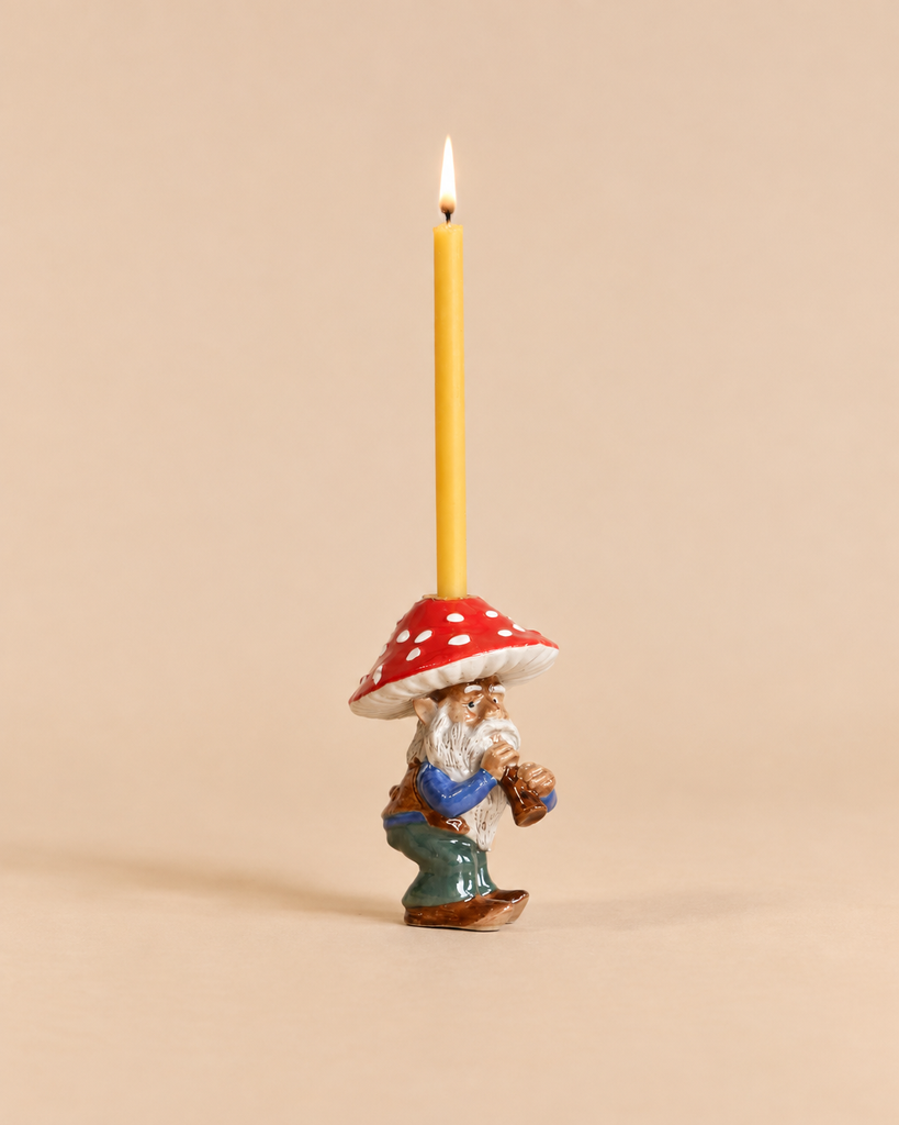 Add a whimsical touch to your space with the Mushroom Gnome Cake Topper—this heirloom-quality collectible features a ceramic gnome holding a lit yellow candle, dressed in a blue shirt and green pants with a red, white-spotted mushroom hat. Perfect as a keepsake or unique decor accent, this final sale item is photographed against a plain beige background.