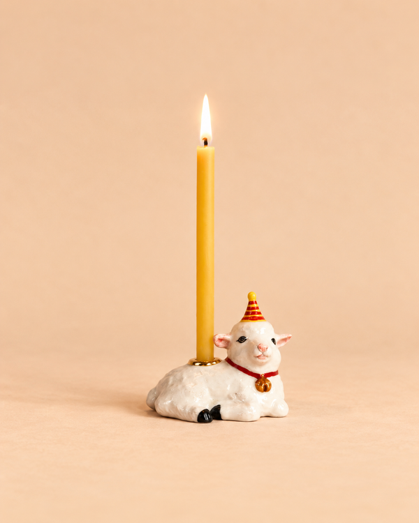 A lit beeswax birthday candle is held by a ceramic goat wearing a red and yellow party hat, set against a plain beige background—an adorable collectible keepsake for any special celebration. The Goat Cake Topper - Final Sale adds charm to your festivities.