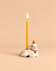 A lit beeswax birthday candle is held by a ceramic goat wearing a red and yellow party hat, set against a plain beige background—an adorable collectible keepsake for any special celebration. The Goat Cake Topper - Final Sale adds charm to your festivities.