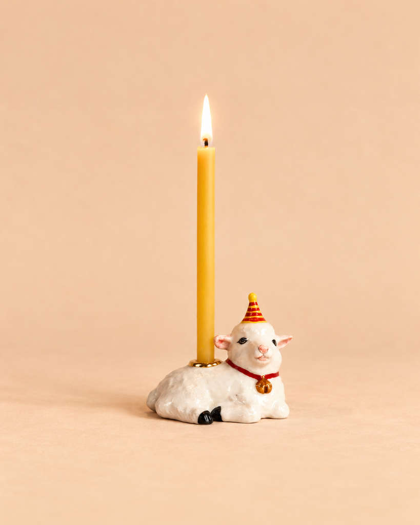 A lit beeswax birthday candle is held by a ceramic goat wearing a red and yellow party hat, set against a plain beige background—an adorable collectible keepsake for any special celebration. The Goat Cake Topper - Final Sale adds charm to your festivities.