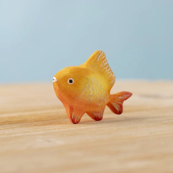 The Bumbu Handmade Wooden Golden Fish, a small orange and yellow Montessori Waldorf toy with painted scales and fins, rests on a light wooden surface against a soft blue background.