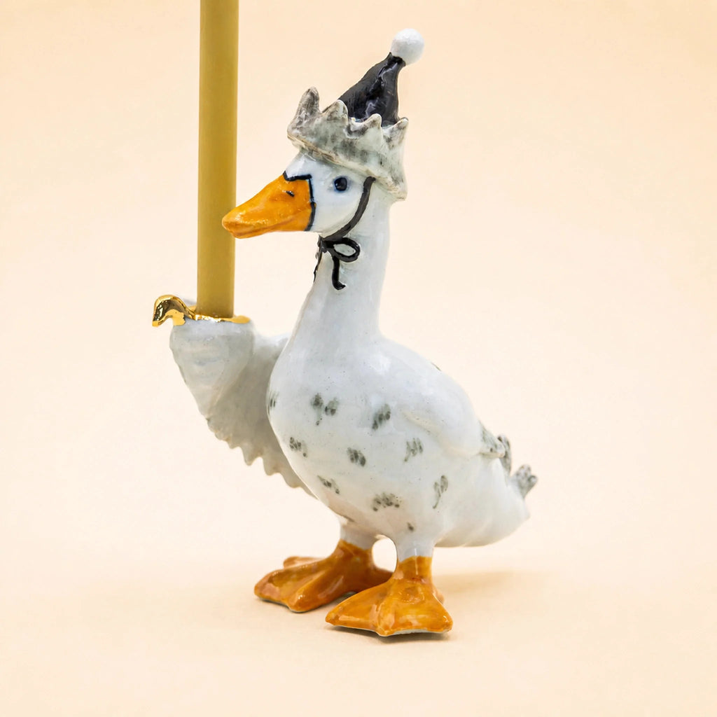 Meet the Goodnight Goose Cake Topper – Final Sale: a charming handcrafted collectible featuring a ceramic goose figurine wearing a festive party hat and holding a thin yellow candle upright with one wing against a simple beige background. Perfect as a keepsake birthday decoration, it adds whimsical delight to any celebration.