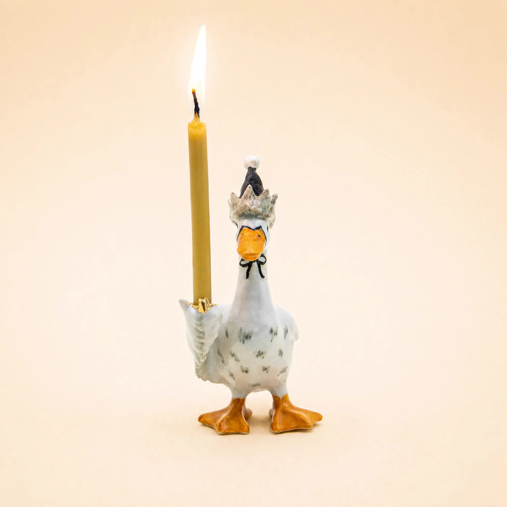 The Goodnight Goose Cake Topper features a charming ceramic goose wearing a small hat and holding an upright lit yellow candle in one wing, set against a plain beige background—an ideal handcrafted keepsake or birthday decoration for any celebration.