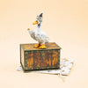 The Goodnight Goose Cake Topper features a charming ceramic goose donning a party hat and holding a gold ring, poised atop a decorative wooden chest set on a star-patterned white cloth against a soft beige backdrop—making it an ideal handcrafted collectible or keepsake birthday decoration.