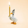 The Goodnight Goose Cake Topper, a handcrafted collectible ceramic figurine featuring orange feet and beak, a gray hat, and an upright lit yellow candle in one wing, is displayed against a pale peach background.