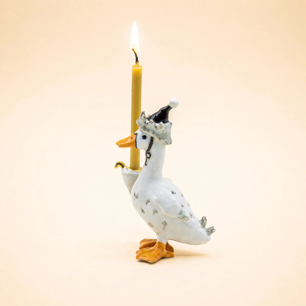 The Goodnight Goose Cake Topper, a handcrafted collectible ceramic figurine featuring a goose wearing a party hat and holding an upright lit candle with one wing, stands out as a charming keepsake birthday decoration against a plain beige background.
