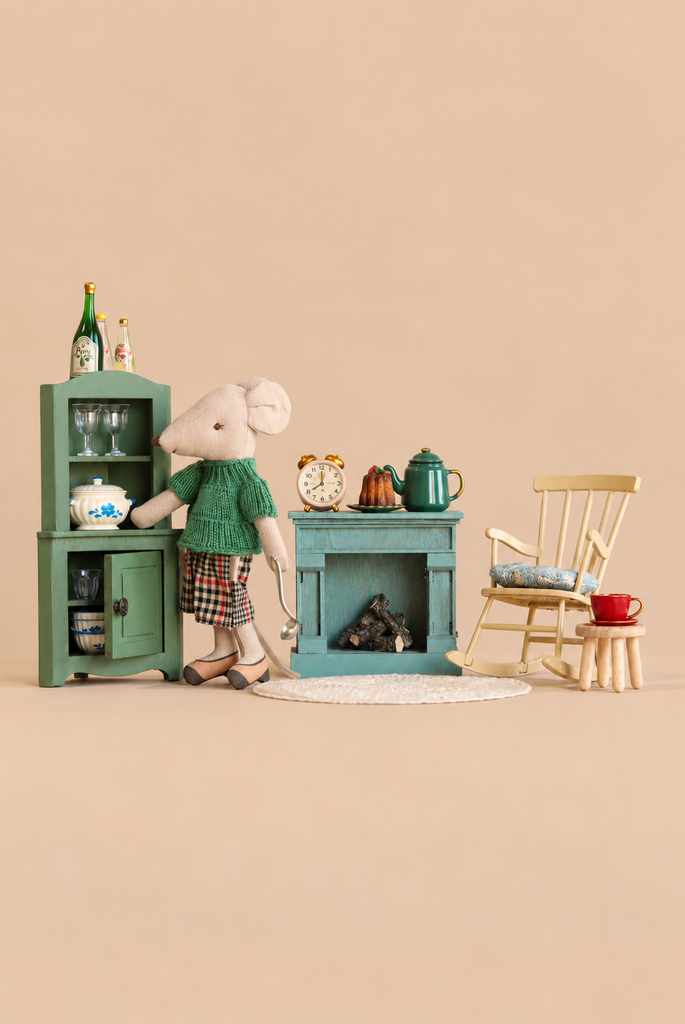 A Maileg mouse doll in a green sweater and plaid skirt stands beside a miniature green hutch, a fireplace with a teapot and cake, and a wooden rocking chair from the Maileg Grandma’s Living Room Set.