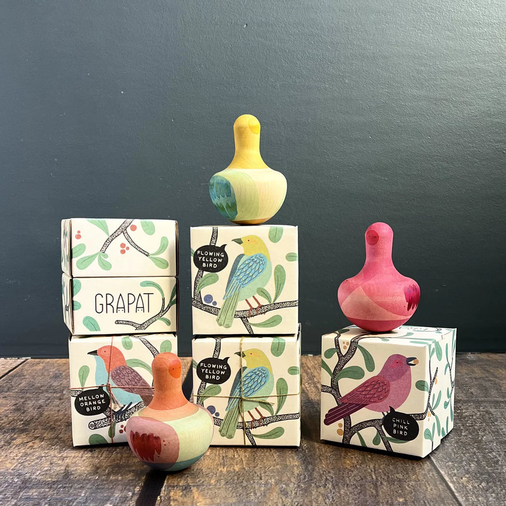 The Grapat Chill Pink Bird Rocking Toy, displayed on illustrated boxes with bird and branch designs, features vibrant wooden toys enhanced by playful feathers. Set against a dark green wall, the wooden floor complements the lively scene. Ships in approximately one week.