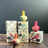 The Grapat Flowing Yellow Bird Rocking Toys, some sporting playful wings, are positioned atop cardboard boxes with bird illustrations. These boxes form two neat columns against a dark green wall, resting on a wooden floor. Ships in approximately one week.