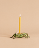 The “Cricket Cake Topper - Final Sale” is a green, hand-painted porcelain candle holder shaped like a grasshopper, featuring a yellow birthday candle on its back. The grasshopper wears a small white party hat adorned with red dots, set against a plain beige background.