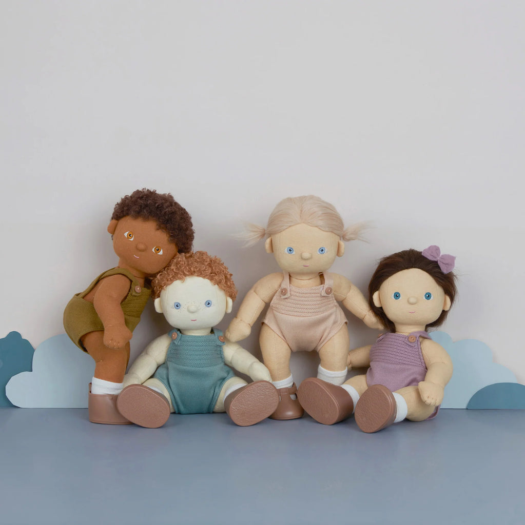 Four diverse Olli Ella | Dinkum Doll - Button with different skin tones and hair colors are sitting against a blue backdrop with white cloud shapes.