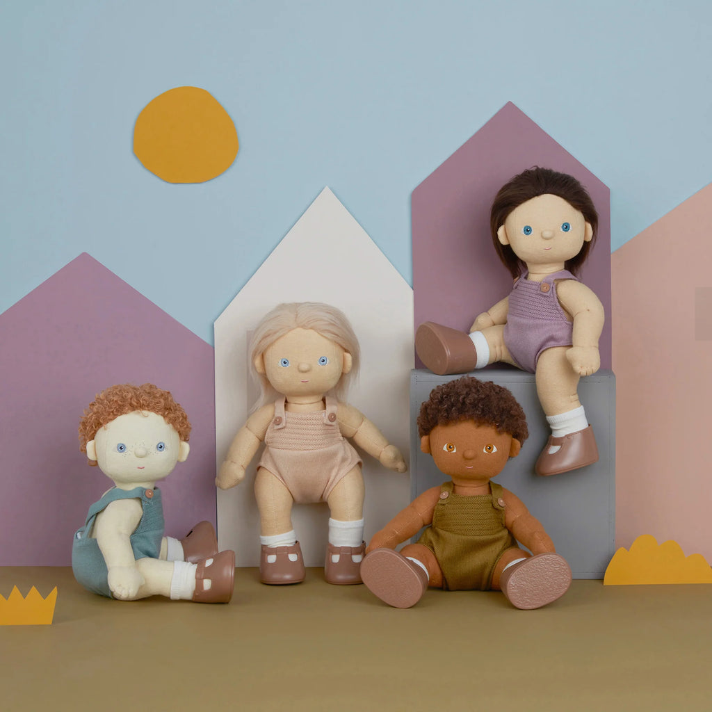 Four diverse Olli Ella | Dinkum Doll - Pea sit against a backdrop with stylized pastel houses and abstract sun shapes, creating a playful and imaginative scene.