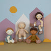 Four diverse Olli Ella | Dinkum Doll - Button seated in front of pastel-colored house-shaped cutouts, with stylized sun and clouds in the background. Each doll has unique hair and outfit colors.