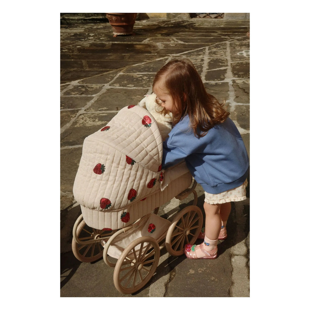 A young girl in a blue jacket and pink sandals smiles as she plays outdoors with the Konges Sloejd Mini Doll Pram - Strawberry, a vintage-style pram with large wheels decorated with red strawberries on stone pavement.