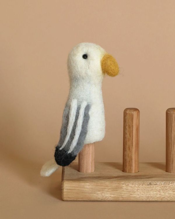 A handmade, needle felted Seagull Finger Puppet, featuring a yellow beak, white body, and grey wings, stands perched on a wooden stand. Made from 100% wool, it sits against a solid, neutral beige background.