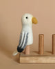 A handmade, needle felted Seagull Finger Puppet, featuring a yellow beak, white body, and grey wings, stands perched on a wooden stand. Made from 100% wool, it sits against a solid, neutral beige background.