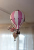 The Hot Air Balloon - Powder Pink is a handmade pink and white striped decoration with a small stuffed animal, perfect for children's rooms. It hangs indoors with sheer curtains, while another balloon ornament appears in the background.