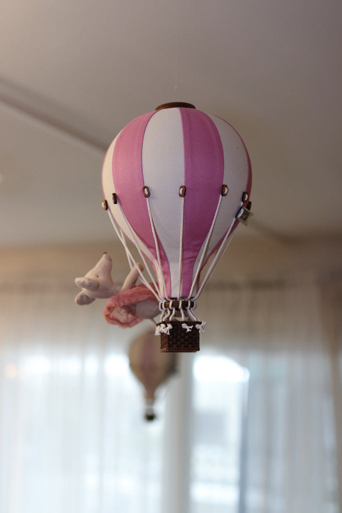 The Hot Air Balloon - Powder Pink is a handmade pink and white striped decoration with a small stuffed animal, perfect for children's rooms. It hangs indoors with sheer curtains, while another balloon ornament appears in the background.