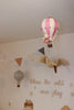 The Hot Air Balloon - Powder Pink hangs from the nursery ceiling, surrounded by flags, a wall-mounted bird, a blue balloon ornament, and the words "Where the wild ones play" on the wall.