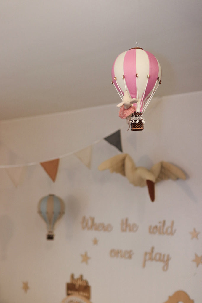 The Hot Air Balloon - Powder Pink hangs from the nursery ceiling, surrounded by flags, a wall-mounted bird, a blue balloon ornament, and the words "Where the wild ones play" on the wall.