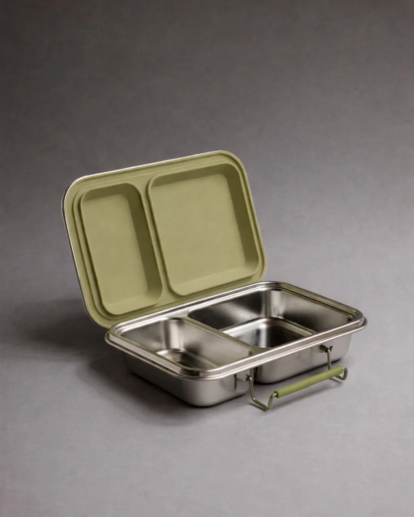 A stainless steel lunch box with two compartments and an olive handle is open, showing its matching olive-colored lid propped up behind it. The Haps Nordic eco-friendly food container is styled as a bento box and placed on a plain grey surface.