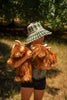 In a sunlit outdoor space where grass meets tree shade, a child in a green and white plaid sun hat holds two Heidi the Highland Cow Stuffed Animals with long brown fur and light horns, enjoying a perfect setting for playful imaginations.