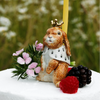 Hand-painted Highness Hare Cake Topper - Final Sale (ships in one week), this ceramic bunny with crown and cape sits on a white cake among pink flowers, berries, and a yellow candle against a green background.