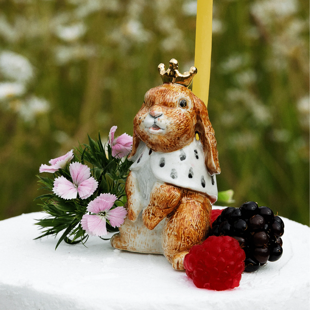 Hand-painted Highness Hare Cake Topper - Final Sale (ships in one week), this ceramic bunny with crown and cape sits on a white cake among pink flowers, berries, and a yellow candle against a green background.