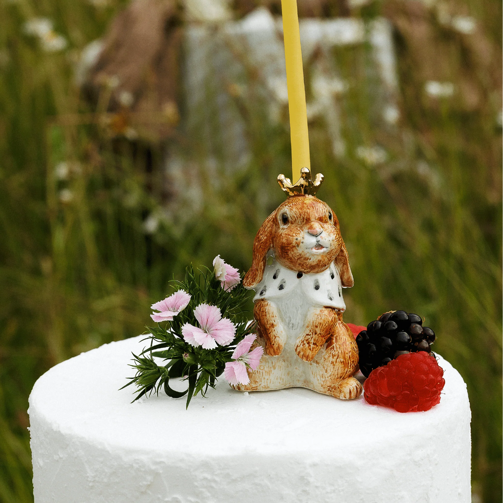 A white cake is adorned with the Highness Hare Cake Topper - Final Sale (ships in one week), fresh flowers, a blackberry, and a raspberry, set against a softly blurred grass and wildflower background.
