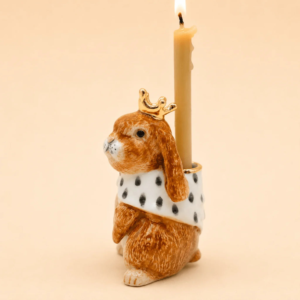 The Highness Hare Cake Topper - Final Sale (ships in one week) features a ceramic bunny with a gold crown and white spotted cape, holding a lit beige candle—a charming porcelain accent for your cake.