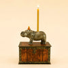 The Hippo Cake Topper - Final Sale features a ceramic hippo wearing a tiny party hat and carrying a lit yellow candle on its back, standing atop a wooden treasure chest with metal accents—a whimsical, handcrafted birthday decoration perfect as a collectible keepsake.