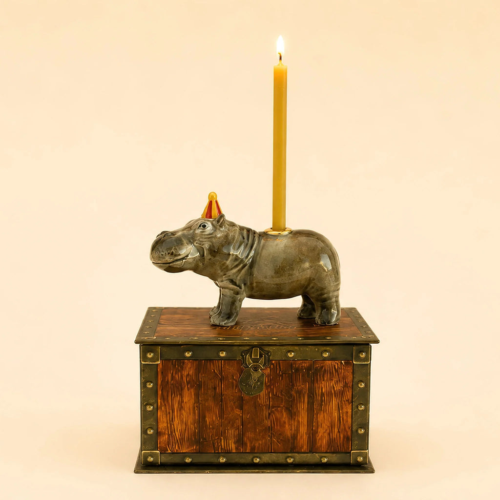 The Hippo Cake Topper - Final Sale features a ceramic hippo wearing a tiny party hat and carrying a lit yellow candle on its back, standing atop a wooden treasure chest with metal accents—a whimsical, handcrafted birthday decoration perfect as a collectible keepsake.