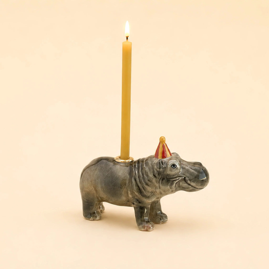 The Hippo Cake Topper - Final Sale features a ceramic hippopotamus figurine wearing a red party hat and holding a lit candle on its back against a pale beige background, making it a charming handcrafted birthday decoration and a delightful collectible keepsake.