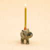 A lit yellow candle sits in the Hippo Cake Topper—a handcrafted ceramic figurine featuring a hippo wearing a small red party hat—set against a plain beige background, making this final sale decoration a charming and collectible keepsake.