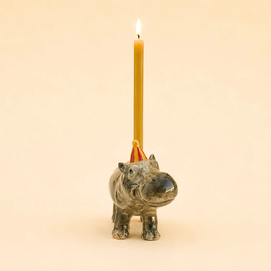 A lit yellow candle sits in the Hippo Cake Topper—a handcrafted ceramic figurine featuring a hippo wearing a small red party hat—set against a plain beige background, making this final sale decoration a charming and collectible keepsake.