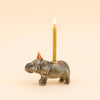 The Hippo Cake Topper - Final Sale features a handcrafted ceramic hippo figurine donning a small red party hat and carrying a lit yellow candle on its back against a plain beige background, making it a delightful birthday decoration and collectible keepsake.