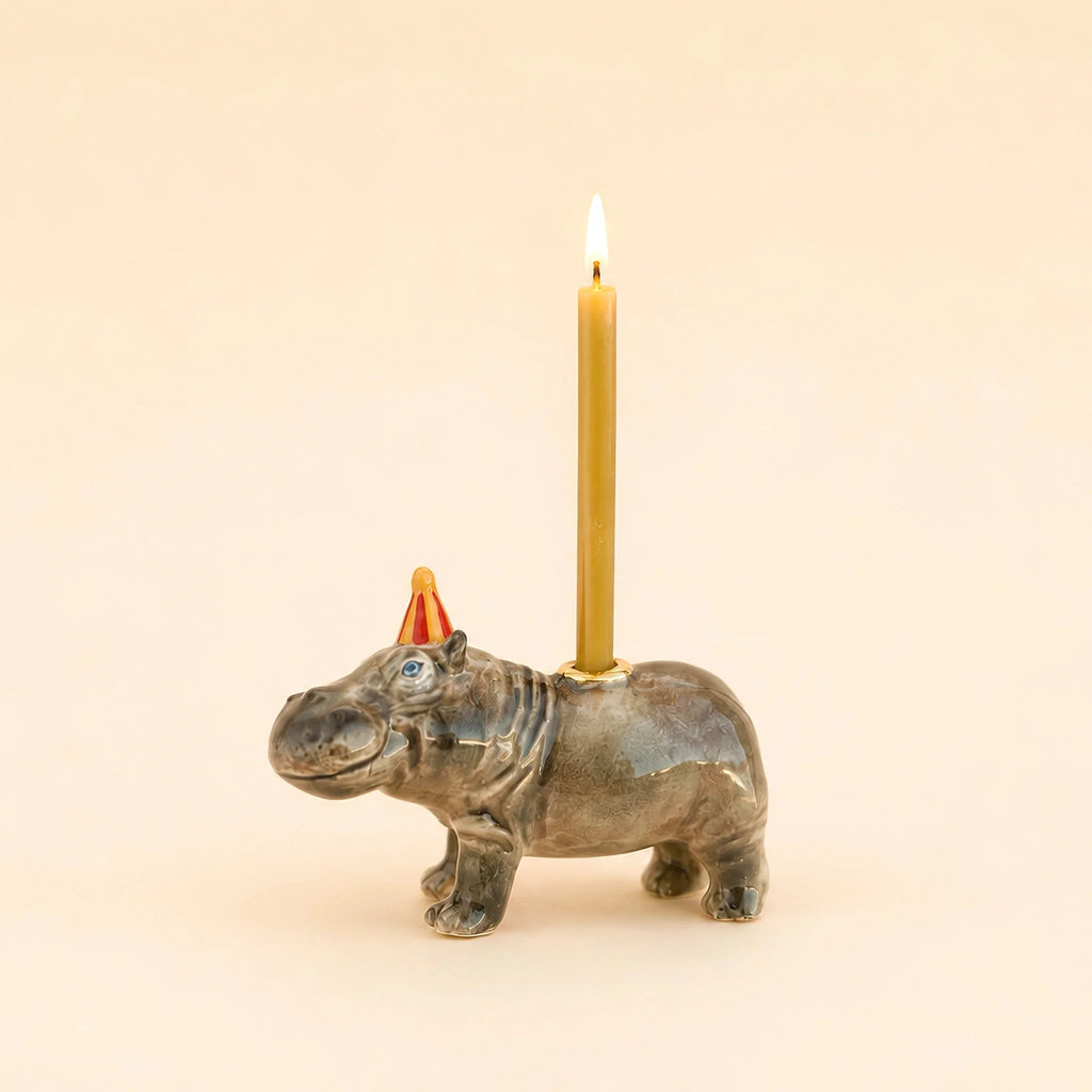 The Hippo Cake Topper - Final Sale features a handcrafted ceramic hippo figurine donning a small red party hat and carrying a lit yellow candle on its back against a plain beige background, making it a delightful birthday decoration and collectible keepsake.