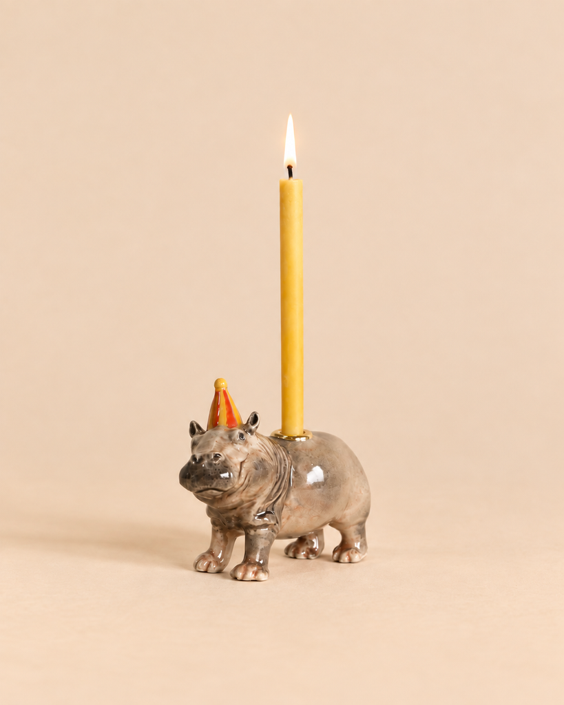 Introducing the Hippo Cake Topper – Final Sale: This handcrafted collectible features a ceramic hippopotamus figurine adorned with a red party hat and carrying a lit yellow candle on its back, set against a plain beige background.