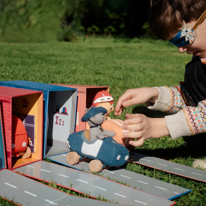 A child in a superhero mask engages in imaginative play outdoors with an Olli Ella Holdie Dog-Go Officer toy and a plush toy, arranging them on a makeshift roadway next to cardboard buildings on grass.