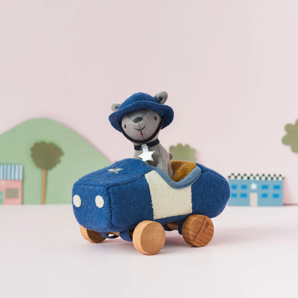 A Olli Ella Holdie Dog-Go Officer plush toy mouse wearing a hat and driving a blue felt car with wooden wheels, set against a pastel backdrop with simple geometric shapes representing trees and houses, perfect for nursery decoration.