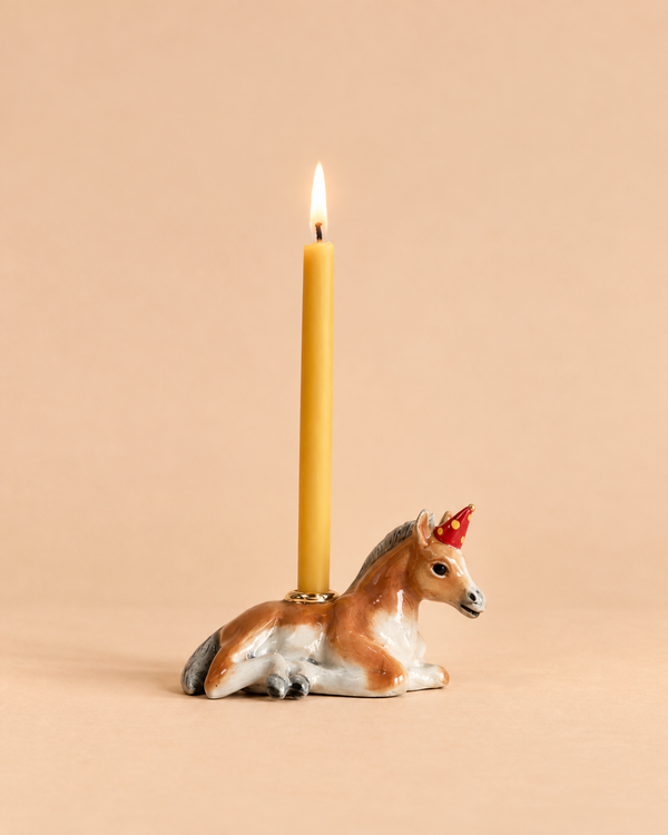 A yellow candle burns in a holder shaped like the “Horse Cake Topper - Final Sale,” which resembles a brown and white unicorn lying down and has the appearance of a handcrafted collectible, set against a plain beige background.
