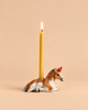 A yellow candle burns in a holder shaped like the “Horse Cake Topper - Final Sale,” which resembles a brown and white unicorn lying down and has the appearance of a handcrafted collectible, set against a plain beige background.