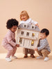 Three young children are smiling and holding the House of Hamptons — Little Forest Animals dollhouse together against a beige background.