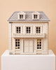 The House of Hamptons — Little Forest Animals, a beige and gray two-story dollhouse featuring a balcony and four dormer windows, is displayed on a white pedestal against a plain tan backdrop, capturing the inviting East Coast style ideal for Little Forest Animals to make their home.